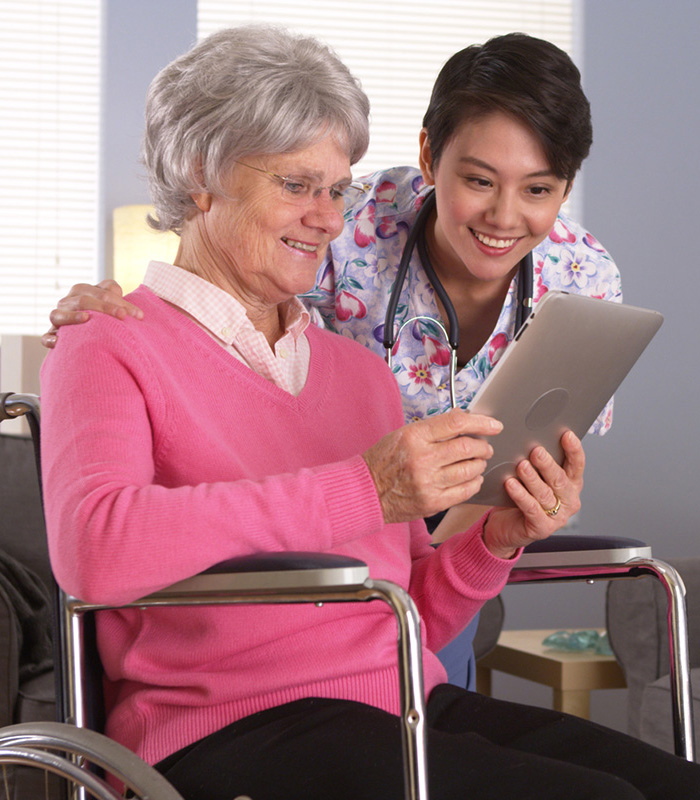 A caregiver with an elderly woman in a wheelchair reviewing info on a tablet computer